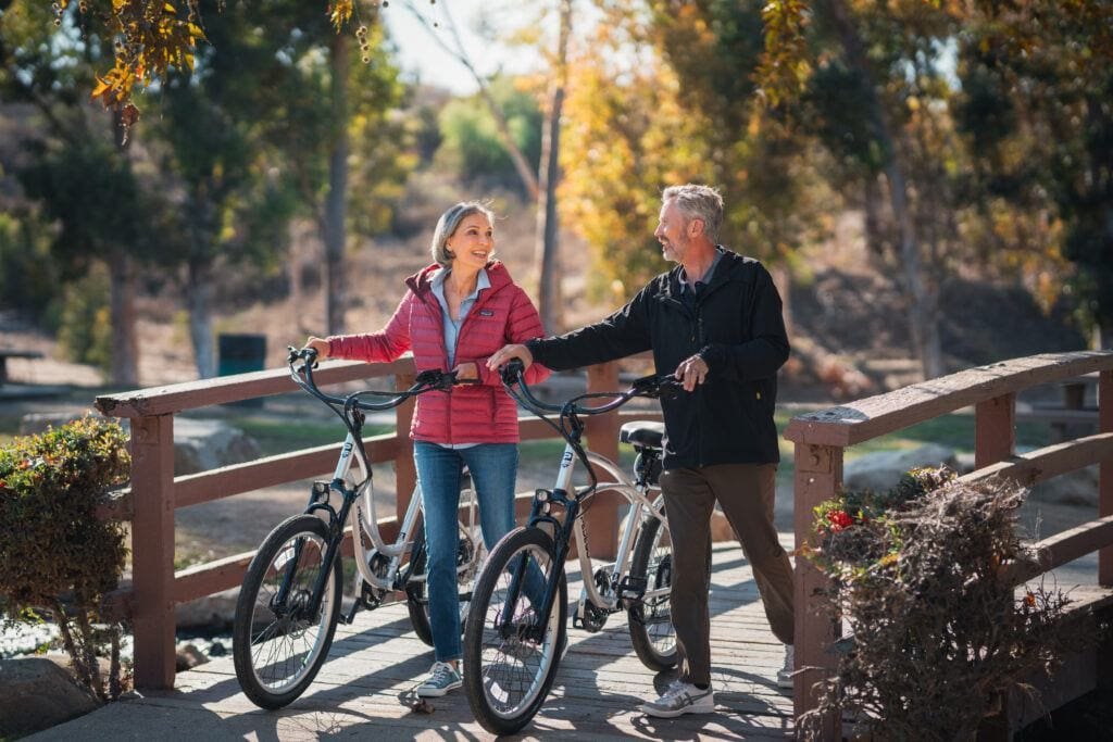 A couple walking through the woods over a bridge with their Pedego Interceptor Electric Bikes. 